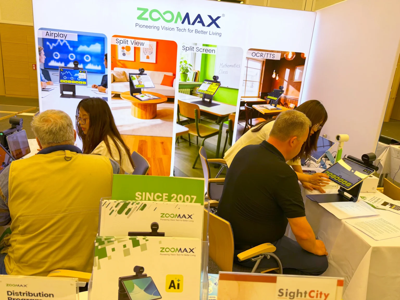 SightCity 2026 fuarındaki Zoomax standında yoğun ziyaretçi trafiği görülüyor. Katılımcılar Snow Pad serisi elektronik büyüteçleri ve Snow Eye Pro cihazını deneyimleyerek ürünler hakkında bilgi alıyor.